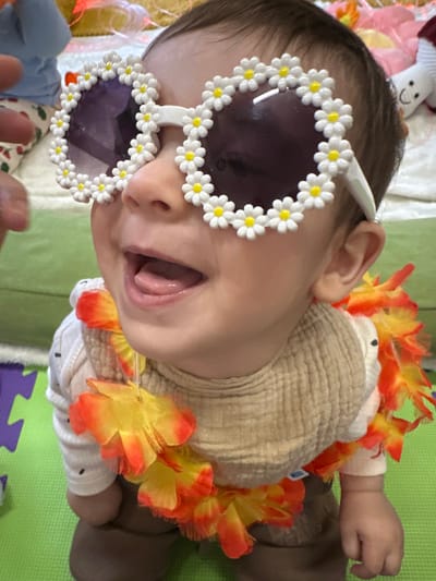 A baby is depicted in an indoor setting in Prestbury, United Kingdom, facing slightly upwards and towards the viewer's right. The baby wears oversized, white-framed sunglasses adorned with white daisy flowers and dark purple tinted lenses. A multi-colored lei, primarily yellow and orange, is draped around the baby's neck. Underneath the lei, a beige, textured bib is visible over a light-colored, long-sleeved top with small dark speckles. The baby's mouth is open, revealing the tongue, indicating a laugh or wide smile. Dark brown trousers are visible on the lower body. A partial view of an adult's hand, including an index finger and thumb, is visible on the left, near the baby's upper arm. The baby is positioned on a light green playmat with a visible purple edge in the bottom left. The background consists of blurred soft furnishings, including a white object resembling a plush toy with a simple black facial feature.