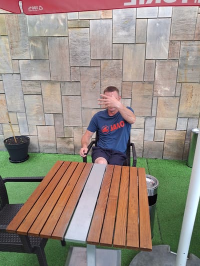 A man sits at a table outdoors, obscuring his face with his hand. He is wearing a dark blue t-shirt with a red logo and lettering that reads "JAKO". He is also wearing dark shorts. The table has a wooden top with a metal inlay. A black plastic pot with a small plant is on the green, artificial turf ground next to the table. Behind the man is a stone wall made of rectangular stone tiles of varying shades of grey and brown. To the right of the man, a metal pole extends upwards and a white pole is visible at the top of the frame. A red banner with white text is visible on the left side of the image. The weather appears to be clear. The lighting suggests it is daytime. The location is Pila, Poland. There is text on the red banner that includes "100%" and a recycling symbol.