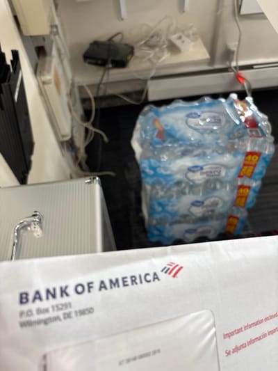 An indoor, medium-shot photograph displays a collection of objects, primarily focused on an envelope and packaged bottled water, against a backdrop of electronic devices and cables. In the right foreground, a white envelope from "BANK OF AMERICA" is prominently visible, oriented vertically. Visible text on the envelope includes "BANK OF AMERICA", "P.O. Box 15291", and "Wilmington, DE 19850". Further up, "Important information enclosed" and "Se adjunta información import." (Spanish for "Important information attached") are also legible.
Behind the envelope and slightly to the left, a silver-colored metallic case with a handle is partially visible. In the upper mid-ground, a multi-pack of bottled water, likely a 24-pack, is stacked. The bottles have blue labels, and red and yellow tags indicating "40" and "VALUE" are attached to the packaging. The bottles appear to be a store brand, possibly "Everyday Essentials" or similar, though text on the bottles is blurry.
To the left of the scene, various electronic components and tangled white and black cables are present, including a black device hanging from what appears to be a shelf or a wall-mounted hook, and a white power adapter. A long, white rectangular device with some orange labeling is also visible on a surface, likely a desk or counter. The background behind the bottled water is dark, suggesting a cabinet or a shadowed area. The overall impression is one of a cluttered utility space or a back office in Hampton, United States of America. No people are visible, and no specific activity is taking place. The lighting suggests indoor artificial illumination. The image has a shallow depth of field, with areas of significant blur.