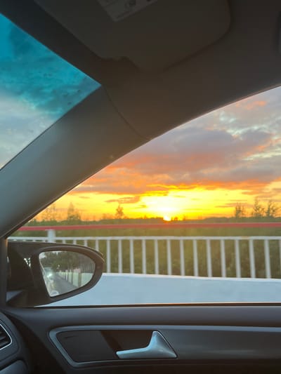 The image presents a view from the interior of a vehicle during sunset in Васильево, Russia. The primary focus is the vibrant sunset observed through the front passenger window. The sky is illuminated with intense yellow and orange hues near the horizon, where the sun is visible as a brilliant, low-lying disc, partially obscured by a dark silhouette of distant trees and vegetation. Above the brighter lower sky, clouds show fiery red, orange, and softer purple tones.
Below the horizon, a structural element, likely part of a bridge or an elevated roadway, is visible. It features a white railing composed of numerous vertical slats and a prominent red horizontal bar running along its length.
The car's interior frames the scene. The upper left corner shows a portion of the windshield with a blue tint, possibly from a panoramic roof or a reflection, and numerous small circular spots on the glass surface. A light gray A-pillar diagonally borders the main view on the left. In the lower left foreground, a black side mirror reflects a road or pathway that runs parallel to a white fence or wall, with a distant light source, possibly a vehicle headlight or streetlamp, visible within the reflection. The lower section of the image shows the car's dark gray door panel, featuring a silver-colored interior door handle and a portion of an air vent on the far left. No people are visible in the image.