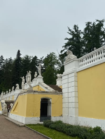 The image depicts a section of the palace in Arkhangel'skoe, Russia. The building is painted yellow with white architectural details, including a balustrade featuring sculpted figures. A window with a dark metal grate is visible. Trees and other greenery are present in the background. The sky is overcast.