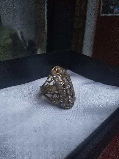 A single ornate ring rests within a black, rectangular container, specifically upon a white, textured insert. The ring exhibits an aged, gold-toned metal base with an elongated, openwork filigree design, possibly featuring a lattice or honeycomb pattern. Its upper surface is densely set with numerous small, dull, clear faceted stones, likely imitation diamonds or crystals. The immediate background outside the container is dark and blurred, showing minimal detail, with a faint reddish hue visible towards the right, consistent with an indoor setting. No persons are present, and no actions are occurring. The location of this scene is the city of Gerasimovskoye, Kazakhstan.