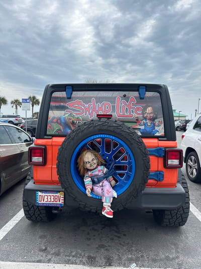 This image shows the back of an orange Jeep Wrangler, parked in a parking lot. The vehicle has custom details: a spare tire with a bright blue rim and a Chucky doll positioned within the tire. The back window has a custom graphic. A Florida license plate is visible, with the text "DV338VE." The license plate also indicates support for the Florida Gators. Cars surround the Jeep, and palm trees are visible in the background. The scene is observed to be in the city of Jacksonville Beach, United States of America.