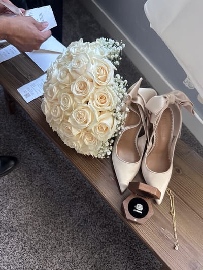 A pair of beige satin high-heeled shoes with bows on the back are placed on a wooden bench. Next to the shoes is a bouquet of white roses and baby's breath. A hexagonal wooden box holding a diamond engagement ring and a gold necklace with a pearl pendant are also on the bench. A person's hands are visible holding a card and an envelope. The scene appears to be indoors with a gray carpeted floor and a white wall in the background. The specific location in El Paso, United States of America cannot be determined from the provided image.