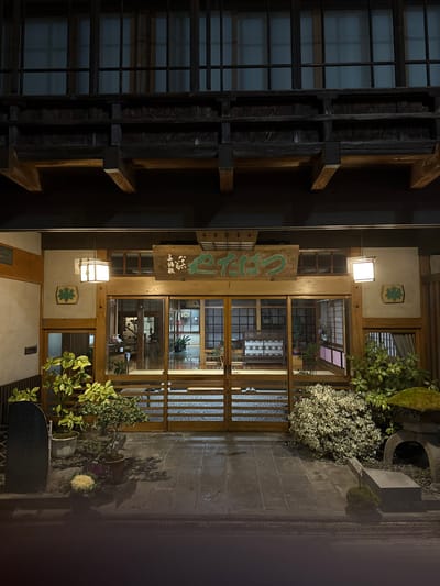 An exterior, nighttime image captures the illuminated entrance of a traditional Japanese building identified as "旅館 こたけつ" (Ryokan Kotaketsu) in Yamanouchi, Japan. The structure features dark wooden beams and an upper level with numerous lattice windows. Below, a large, rectangular wooden sign with green Japanese characters is centered above the main entrance. Two square, white-glowing lanterns flank this sign and the entrance doors. The entrance comprises multiple paneled glass and wooden sliding doors, through which the warmly lit interior with tiled flooring and potted plants is visible. A green maple leaf emblem is affixed to the wall on the left of the entrance.

The ground area immediately in front of the building consists of dark, wet-looking stone paving. To the left, a tall, dark stone monument stands alongside several potted plants, including a variegated leafy plant and a smaller bonsai. To the right, a dense, variegated bush with light-colored foliage is present next to a traditional stone lantern topped with moss and smaller mossy mounds. No individuals are visible within the frame. The overall lighting is artificial, emanating from the building's interior and exterior lanterns, contrasting with the dark surrounding environment.