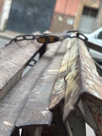 This is a close-up, low-angle shot of a rusty metal structure, possibly a bench or a part of a vehicle. The metal is weathered, showing patches of rust and old paint in shades of brown, yellow, and some greenish-blue. A metal chain is draped across the top of the structure, secured with a small, brass-colored padlock. The background is blurred, showing elements of an urban environment. On the left, a dark, textured surface is visible, possibly a wall or a door. To the right, the blurred shapes of a building with windows and a white vehicle suggest a street or a parking area. The overall impression is one of disuse and decay. This scene is likely located in Berrechid, Morocco, given the context of its origin, suggesting an outdoor setting within the city.