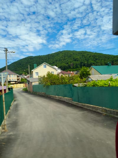 The image shows a street scene in Agoy, Russia, on a cloudy day. A paved road curves to the left, bordered by a green fence on the right. Several houses with yellow and green roofs are visible behind the fence. A lush green hill rises in the background, covered in trees. The sky is blue with scattered white clouds.