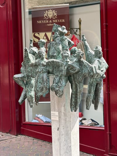 This is a medium shot, eye-level, outdoor, eye-level photograph of a bronze sculpture of a group of people on a concrete pedestal, located in front of a building with a storefront in Roermond, Netherlands. The sculpture, which depicts a cluster of figures with abstracted features and stylized clothing, appears to be in motion, with some figures raising their arms and others seemingly marching or dancing. The bronze has a weathered, greenish patina, suggesting age and exposure to the elements.

The building behind the sculpture has a large glass window displaying clothing, with visible price tags. A sign on the window reads "MEYER & MEYR - tailors," and another sign below it says "SALE". The storefront is painted a vibrant red. The sidewalk in front of the building is paved with red brick.

A small, laminated plaque is attached to the concrete pedestal. It is partially obscured by the sculpture but appears to have text written in Dutch. Through the glass, some clothing items are visible in the storefront window, including folded shirts with patterns and some sweaters. The general impression is that of a public art installation in a commercial area. The sculpture's name is "De Ruivers" and it was created by Theo van de Logt in 1992.