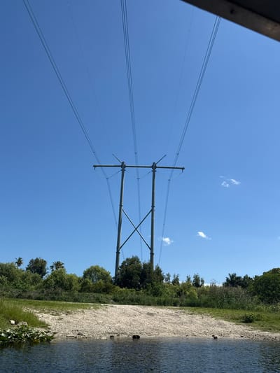 The image shows a landscape view with a focus on power lines and the sky. A large metal power pole dominates the scene, stretching upwards against a clear blue sky dotted with a few small clouds. Power lines extend from the pole across the frame.
To the right of the power pole, there is a grassy bank along the edge of a body of water (likely a canal or river). The shoreline is lined with vegetation, including trees and shrubs. The water appears calm.
It is not possible to confirm the precise location of the image in Fort Lauderdale based solely on visual information.