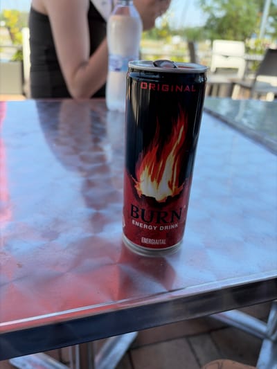 A close-up shot focuses on a red and black can of "Burn Energy Drink" placed on a reflective metallic table. The can features a design of orange and yellow flames. The word "ORIGINAL" is visible in red lettering at the top of the can, and "BURN ENERGY DRINK" is also clearly legible. Below that, the word "ENERGIAITAL" is present. In the background, slightly out of focus, a person wearing a black tank top is visible, along with a partially obscured plastic bottle of water. The setting appears to be an outdoor area, possibly a cafe or restaurant, with trees and outdoor furniture in the distant background. The metallic surface of the table reflects the environment, creating a patterned effect. The context of Mogyoród, Hungary, suggests this scene could be from a location in or around the Mogyoród Formula 1 circuit or a nearby establishment.