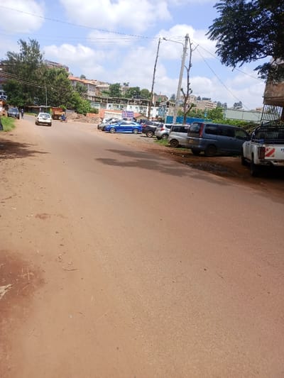 The image presents a daytime street scene in Murang'a, Kenya. A reddish-brown asphalt road, flanked by unpaved earth verges, extends into the background under a partially cloudy sky. On the left side of the road, a white sedan and a motorcycle carrying a rider are observed moving away from the viewer. A lone pedestrian in dark clothing walks further along the left roadside. Dense green foliage and trees line this side of the road, with multi-story buildings visible on an elevated terrain in the distance. On the right side of the road, a series of approximately seven vehicles is parked at an angle. These include a white Isuzu pickup truck with its rear facing the viewer in the immediate foreground, followed by a dark grey minivan, a silver hatchback, a dark grey SUV, and a blue sedan. A utility pole with power lines stands vertically on the right. In the middle ground, beyond the parked vehicles, additional buildings, some with corrugated iron roofs, are visible on a hill, along with a prominent blue and white sign displaying partial text such as "K-MART" and "ELECTRONICS."