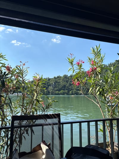 The image depicts a serene lakeside view from a covered outdoor seating area. In the foreground, a black metal chair with a textured seat is positioned near a black metal railing. A cream-colored bag with brown accents rests on the chair. Flowering plants with pink blossoms and green leaves are in bloom, framing the view.
Beyond the railing, a calm, green body of water stretches out, reflecting the clear blue sky dotted with white clouds. In the middle ground, a lush, tree-covered hillside rises steeply from the water's edge. A few small boats are visible on the lake. In the distance, a red-roofed building can be seen nestled on the hillside.
The scene is set in Divic, Bosnia and Herzegovina, suggesting a location with natural beauty and recreational opportunities. The overall atmosphere is peaceful and inviting, characteristic of a lakeside resort or restaurant. The time of day appears to be daytime, with bright sunlight casting shadows.
