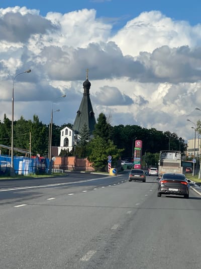 The image shows a street scene in Bittsa, Russia. On the left side of the road is an Orthodox church with a black dome and gold cross, partially obscured by trees. A large, cloudy sky dominates the background. The right side of the image is dominated by a gray asphalt road with white lane markings. There are several vehicles on the road, including cars and a truck. A gas station sign, labeled "AVIS", is visible amidst green foliage. The scene suggests a mix of urban and religious elements in the Bittsa landscape. Construction materials are present near the church.