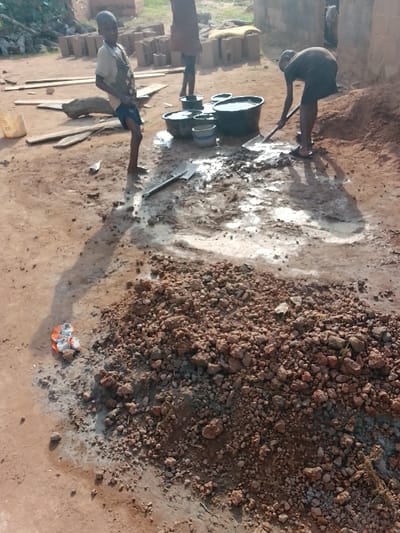 The image captures an outdoor construction or brick-making site in Ugwolawo, Nigeria, under daylight conditions. The ground is unpaved, consisting of reddish-brown dirt, with several areas covered in a whitish-grey liquid mixture.

Three individuals are present. On the left, a young boy with short hair, wearing a light-colored top and dark shorts, stands upright, facing slightly right. In the center-rear, another individual, partially obscured from the waist down, stands near a cluster of containers. To the right, an adult or older youth, dressed in a dark top and dark shorts, is actively bending over and using a shovel to mix the whitish-grey substance on the ground. This individual wears flip-flops.

Essential objects include:
- A pile of reddish-brown granular material, likely soil or aggregate, occupying a significant portion of the lower right foreground.
- Four dark-colored buckets, some filled with water, and one smaller light-colored bowl, clustered together in the mid-ground.
- Two shovels: one in active use by the right individual, and another lying flat on the ground.
- Stacks of numerous rectangular blocks, possibly unbaked bricks or building materials, visible in the background.
- Several wooden planks scattered on the ground to the left.
- A crumpled orange and silver object resting on the ground in the lower left foreground.

Long shadows are cast on the ground, indicating direct sunlight.