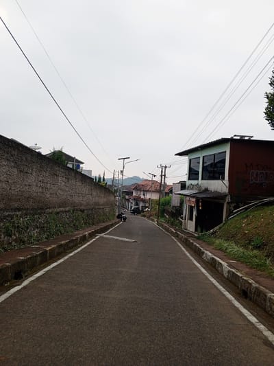 The image depicts a street scene in Pelwehera, Sri Lanka. The perspective is from a low angle looking up a sloping road. On the left side of the road is a tall, weathered concrete wall with a satellite dish visible at the top. Vegetation grows at the base of the wall. On the right side of the road, there is a two-story building with a green upper floor featuring large windows and a red-brown lower section with graffiti on its side. Below this building is a smaller structure with a dark entrance. A motorcycle is parked on the side of the road in the middle distance. Further up the road, several houses with tiled roofs are visible. Electrical wires stretch across the overcast sky. The road itself is paved and has white lines marking the edges. The overall impression is of a residential street in a somewhat hilly or elevated area.
