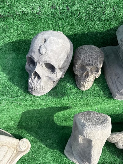 The image depicts a collection of stone skull sculptures, along with other architectural fragments, lying on a vibrant green surface resembling artificial grass. The scene is brightly lit, casting distinct shadows from the objects onto the green surface. The skulls vary in design; one has intricate patterns carved into its surface, while the others are more plain. The other objects appear to be architectural elements, such as pieces of columns or capitals. The image provides no information about whether this is in Chirk or anywhere else.
