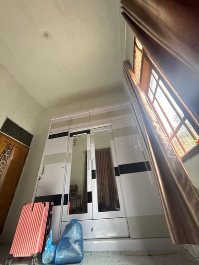 A wide-angle interior shot captures a room with a large white wardrobe, a pink suitcase, and blue trash bags. The wardrobe has mirrored doors and is accented with beige and black stripes. A brown curtain is drawn across a wooden-framed window, allowing sunlight to stream in. The room appears to be a bedroom or storage area in a residential building. The overall impression is one of a functional living space.