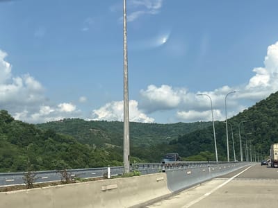 The image captures a view from a highway overpass in Kveda Tsiplavake, Georgia, on a bright, partly cloudy day. Lush, green, forested hillsides dominate the background, with scattered buildings visible on the distant slopes. The foreground features a concrete barrier and a road with multiple lanes. Streetlights line the right side of the highway, and a tall metal pole stands prominently in the center-left of the frame. Several vehicles, including cars and a truck, are present on the road, moving in the same direction. The sky is a clear blue with fluffy white clouds.