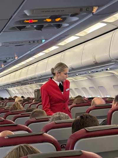 The scene depicts the interior of a commercial airplane cabin, located in Novogorelovo, Russia. A blonde female flight attendant, with her hair styled in a bun, stands in the aisle. She wears a red uniform blazer over a white collared shirt and a black tie, with a nametag visible on her left lapel. Her gaze is directed towards her right. Multiple rows of passenger seats, upholstered in maroon and gray, extend into the background. Numerous passengers are seated, primarily visible from the back of their heads and shoulders, exhibiting varied hair colors including blonde, brown, gray, and bald. Overhead bins line the cabin ceiling, which is illuminated by internal strip lighting. Passenger service units are visible above the seats, featuring individual lights and illuminated red "fasten seatbelt" symbols. The cabin walls are white. No specific interactions are visible between the flight attendant and passengers; she appears to be in motion or surveying the cabin.