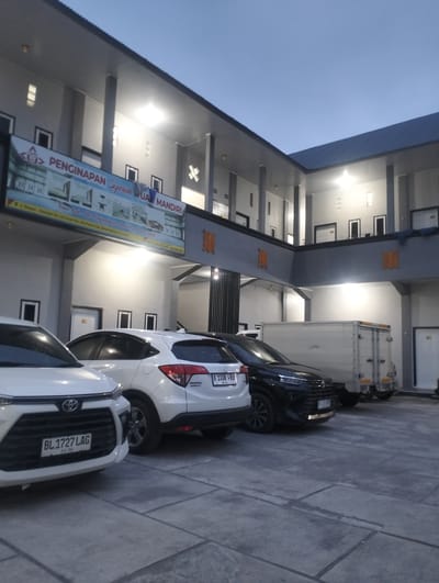 This media file depicts the exterior of a building, likely a lodging establishment, in Panteraya, Indonesia. The building features a modern architectural style with multiple levels, covered walkways, and large windows. The scene is illuminated by artificial lighting, suggesting it is either dusk or nighttime.

In the foreground, several vehicles are parked on a paved surface. These include a white car with the license plate "BL 1727 LAG", a white SUV with the license plate "B 1338 FRU", and a black car. Behind the cars, a white box truck is visible. The building itself has a clean, gray facade with dark gray accents on the walkways and roofline. Some windows are illuminated from within.

A prominent banner is displayed on the front of the building, displaying text in Indonesian and images of rooms. The text is partially obscured but includes "PENGINAPAN Syariah QUAL MANDIRI". This translates to "Syariah QUAL MANDIRI Inn". This banner indicates the nature of the establishment. Orange decorative elements are mounted on the exterior walls. The overall mood is calm and orderly. The sky above is a clear, dark blue, consistent with the evening or early morning.