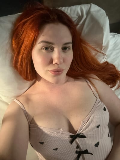 A close-up, high-angle shot shows a woman with vibrant red hair lying on a white pillow. She is wearing a light pink camisole adorned with black heart patterns and small black bows. The woman's eyes are open and looking directly at the camera. The background is a white surface, likely a bedsheet. The setting appears to be a bedroom. There is no discernible indication of the city of Avcılar, Turkey, within the image itself.