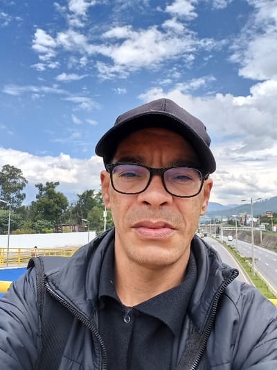 A selfie shows a man wearing a black cap and glasses. He is wearing a dark jacket or coat over a black shirt. The background features a blue sky with scattered white clouds. Also visible are trees and a highway with a yellow barrier, suggesting an elevated position. The perspective is tilted, giving a slightly disorienting view of the sky and landscape behind him.