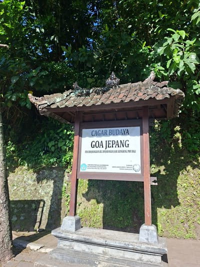 In Banjarangkan, Indonesia, a cultural heritage site sign stands under a traditional tiled roof, supported by wooden posts. The sign, primarily white with dark text, reads "CAGAR BUDAYA GOA JEPANG" and further specifies "DESA BANJARANGKAN, KEC. BANJARANGKAN, KAB. KLUNGKUNG, PROV. BALI". Below this, smaller text details government ministries and regulations. The structure is situated outdoors, with lush green trees and foliage forming a dense backdrop. To the left, a portion of a tree trunk is visible. The ground in front of the sign is paved with stone tiles, and a textured stone wall covered in moss and small plants is behind it. Sunlight casts shadows, indicating a bright, clear day. The scene is static, with no people or discernible activity.