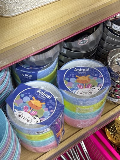 The image shows a shelf in a store in Kebumen, Indonesia, filled with stacks of colorful plastic bowls and metal bowls. In the foreground, there are several stacks of plastic bowls wrapped in clear plastic. Each stack is shrink-wrapped, and the top bowls have blue labels with cartoon animal designs. One label reads "Animal Paradise" and features a giraffe and pandas. Another label states "4 PCS" and "卡通·碗". The bowls themselves are in pastel colors like pink, blue, yellow, and green.

Behind the plastic bowls, there are stacks of metal bowls. Some of these metal bowls have labels attached to them, indicating prices and product information. The shelf itself appears to be made of wood, with a reddish-pink edge. The lighting suggests an indoor retail environment. In the bottom right corner, a pink utensil organizer filled with silverware and a gold-colored strainer are visible. The scene is a close-up view of merchandise on a retail shelf.
