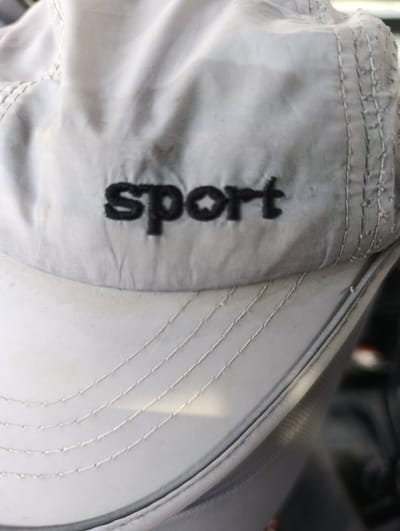 The image shows a close-up of a light gray baseball cap. The word "sport" is embroidered in black, bold letters across the front of the cap. The fabric of the cap appears slightly worn and has a few faint stains. The cap is positioned at an angle, with the brim facing forward. The background is blurred and dark, suggesting it might be indoors or in a dimly lit environment. There is no indication of the location in Khuroson, Tajikistan.