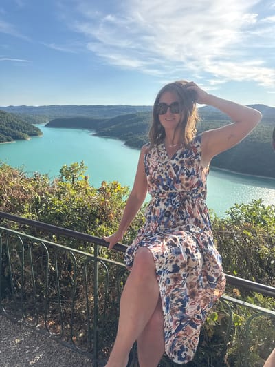 A woman is seated on a railing in front of a panoramic view of a turquoise lake surrounded by verdant hills. She is wearing a floral print dress and sunglasses, with her left hand resting on her head. The sun is shining brightly, casting shadows and highlighting the textures of the foliage and the water. The railing she is sitting on is made of dark metal with decorative curves. The background shows a vast expanse of water winding through the landscape, with a clear blue sky above. The image was taken in Moirans-en-Montagne, France, likely at a viewpoint overlooking the Lac de Vouglans.