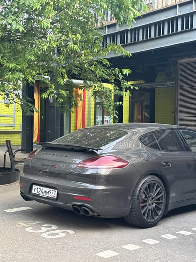 A dark gray Porsche Panamera GTS is parked on a city street. The car is facing to the left, with its rear visible to the viewer. A license plate is mounted on the rear of the vehicle, displaying the characters "Y124MM977" along with "RUS" to indicate its Russian origin. The car has dual exhaust pipes and a spoiler on the trunk. The street is marked with white lines, including a large white "36" painted on the asphalt in front of the car, indicating a parking spot number. A tree with green leaves partially overhangs the car and the building in the background. The building is painted bright yellow with red and yellow banners hanging from its facade. A metal structure with vertical bars is visible above the building's entrance. The scene appears to be outdoors in an urban environment.