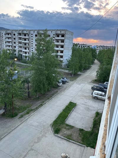 This is an outdoor, medium-angle shot of an abandoned Soviet-era residential complex in Sinegorye, Russia. The sky is overcast with dramatic clouds. In the foreground, a concrete road curves to the left, with a strip of grass to its right. Several vehicles are parked on the road, including a white van, a silver SUV, a dark SUV, and a black SUV. In the middle ground, a tall, unfinished concrete building with many dark, empty window frames dominates the scene. Trees with green foliage are scattered throughout the complex. In the background, more similar buildings and trees are visible, extending into the distance. A dirt road is visible to the right of the middle ground building. The overall impression is one of decay and abandonment.