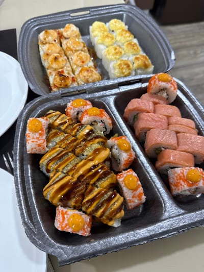 The image shows a close-up, overhead view of two black takeout containers filled with sushi. The containers are divided into compartments. The top container holds two rows of sushi. The left row contains two pieces of sushi topped with a creamy, golden-brown sauce and a drizzle of dark sauce. The right row contains six pieces of sushi topped with a creamy, yellow sauce.

The bottom container is also divided. The left compartment contains several pieces of sushi. The frontmost pieces are topped with a creamy orange sauce and some have orange roe or sesame seeds on the outside. Behind these, there are pieces of sushi with a crispy coating and a drizzle of creamy orange and dark sauce. The right compartment of the bottom container holds rows of sushi rolls with a pink topping, likely salmon, and some have green slivers visible inside. On top of these rolls, there are a few pieces of sushi with orange roe and a dollop of orange sauce.

The scene appears to be indoors, likely on a table, with a hint of a white plate and a fork visible to the left of the containers. The lighting is warm and focused on the sushi. The background is slightly blurred, suggesting a shallow depth of field. There are no people visible in the image. This scene is not specifically located in Ishimbay, Russia, as there are no identifiable landmarks or contextual clues to support such a localization.