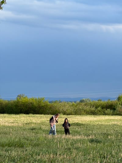 This outdoor image, taken in Jastrebarsko, Croatia, captures a wide-angle view of a natural landscape on a clear, sunny day. The scene is dominated by a large expanse of vibrant blue sky, which occupies the upper half of the frame, featuring a few wispy white clouds. Several thin, parallel power lines are visible horizontally across the sky, just above the horizon.
The lower half of the image consists of a vast, gently undulating field covered in green and yellowish-green grass, suggesting a mix of taller and shorter vegetation or different growth patterns. This field is bordered in the mid-ground by a dense, dark green line of bushes or small trees, creating a natural separation before the distant horizon. Beyond this foliage, a faint, hazy blue line suggests the presence of distant hills or mountains.
Two individuals are present in the grassy field. Closer to the left foreground, one person is standing, dressed in a light-colored, possibly pinkish-brown, jacket over a dark top, blue jeans, and dark shoes. They appear to be looking towards the right side of the frame. Further back and to the right within the field, a second person is reclining or lying down in the grass, wearing dark clothing, including what appears to be a hooded top.
The overall impression is one of a peaceful, leisurely outdoor activity in a rural or semi-rural setting, likely during the late morning or afternoon given the bright sunlight. There is no visible text in the image.