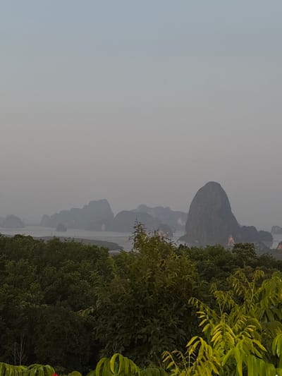 The image shows a hazy landscape with limestone karst formations emerging from a body of water, partially obscured by trees and foliage. The scene appears to be located in Khlong Khian, Thailand, based on the landscape similarities with the city. The sky is overcast. There are no people visible in the image.