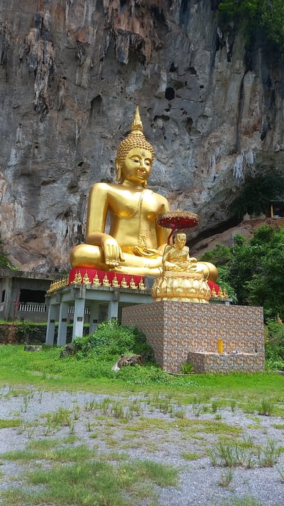 The image depicts a large, golden Buddha statue in a seated pose, situated against a backdrop of a rocky cliff face and verdant vegetation. The statue has a red decorative trim and a smaller figure behind it, possibly another deity. A tiled structure is positioned to the right of the statue. The statue appears to be located outdoors in a landscaped area with gravel and grass. The location is Thap Put, Thailand.
