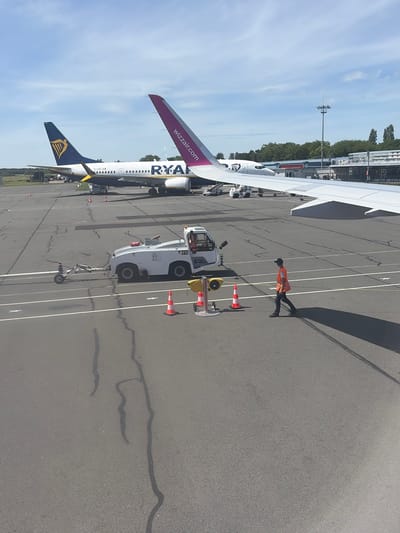 The image captures an airport scene in Tillé, France, featuring two commercial airplanes, one a Ryanair and the other a Wizzair, parked on the tarmac. A tow truck is positioned near the Ryanair plane, suggesting an imminent or ongoing aircraft handling procedure. An individual wearing a high-visibility vest walks near the tow truck and the aircraft, seemingly involved in ground operations. The background shows airport infrastructure, trees, and a blue sky with white clouds. The tarmac is marked with white lines. Traffic cones are also visible, likely placed for safety or guidance purposes.