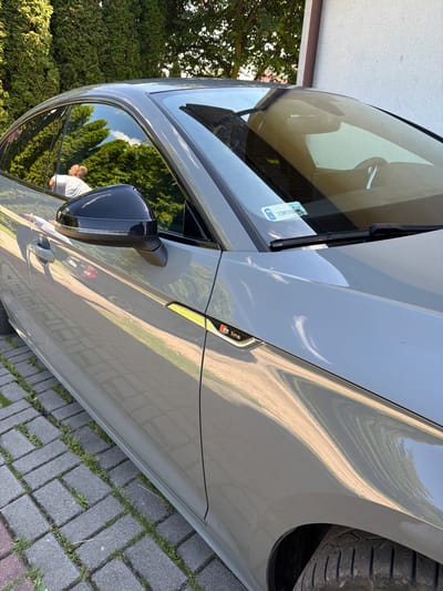 A gray Audi S5 Sportback is parked on a paved driveway in Strzyżowice, Poland. The car's reflection in the side mirror shows a person wearing a white t-shirt, and the car's body is reflecting trees and sky. The car has an "S line" badge on the fender. The driveway is made of interlocking pavers with grass growing between them. A white building is visible in the background to the right of the car.