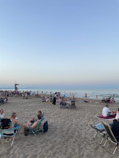 This media file depicts a crowded beach scene in Canet d'en Berenguer, Spain, likely during the late afternoon or early evening. The sky is a soft gradient of blue and pale yellow, suggesting the sun is low on the horizon. The beach is covered in sand, with numerous people scattered across it, many seated in beach chairs or lying on towels.
The subjects are primarily beachgoers enjoying a leisurely day. They are dressed in casual summer attire, including swimwear and t-shirts. Various objects are visible, such as beach chairs, umbrellas, bags, and a lifeguard tower. The scene is one of relaxation and social gathering, with individuals and groups of people interacting or simply enjoying the environment. Some people are in the water, indicating swimming or wading.
Notable details include the sheer number of people, suggesting a popular day at the beach. The presence of a lifeguard tower points to a maintained and likely safe public beach. The overall atmosphere appears relaxed and pleasant, characteristic of a seaside vacation or outing. There is a visible sign on the lifeguard tower which says "Servicio de..." followed by illegible text.