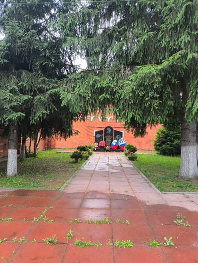 The image depicts a memorial site located in the city of Ivlevo, Russia. In the foreground, a red-tiled pathway, wet from rain and partially overgrown with small green weeds, leads towards the memorial. The pathway is bordered by a grassy area and large evergreen trees with dense foliage on either side. In the midground, a brick wall forms the backdrop of the memorial, which consists of a dark monument featuring carved images and inscriptions, including the dates "1941-1945". Two large floral wreaths, one red, white, and blue, and another with similar colors, are placed at the base of the monument, alongside clusters of colorful flowers. A small, manicured evergreen bush is also visible near the monument. The overall environment is overcast, suggesting a damp, possibly cool, day. There are no people visible in the scene.