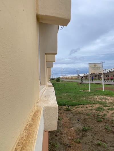 The image depicts an outdoor scene in Dumnga, Nigeria. In the foreground, a beige wall of a building is visible on the left side, extending to the right and showing architectural details like protruding sections and a ledge. The wall has a textured surface and appears to be well-maintained.
Beyond the wall, a grassy area is present, with some bare soil patches. In the mid-ground, a road stretches into the distance, lined with several modest, low-rise buildings that appear to be residential or institutional structures. A signpost stands on the right side of the road, displaying text and a logo, which reads "2016 NATIONAL HOUSING ESTATE" and provides a phone number. Several people are seen in the vicinity of the buildings and the signpost. Some are walking, while others appear to be standing or engaged in activities. Further down the road, more buildings and utility poles are visible, suggesting a developed, though perhaps not densely populated, area.
The sky is overcast with heavy clouds, indicating a cloudy day. The overall environment suggests a semi-urban or suburban setting with new construction or housing development. The scene is captured from a low-angle perspective, emphasizing the building in the foreground and creating a sense of depth as the view extends towards the background.