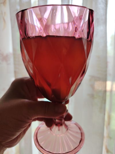 A hand holds a pink, faceted glass goblet filled with a red liquid. The goblet is angled upwards, and the liquid fills about two-thirds of its volume. The background consists of sheer curtains, illuminated by natural light, suggesting an indoor setting. The scene is devoid of any specific landmarks or architectural elements that would indicate a location in the city of Кривец, Russia.