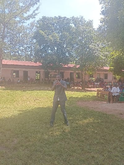 An outdoor scene in Idakho South ward, Kenya, features a male subject in a dark suit playing a dark wind instrument in the foreground of a grassy area. His hands are positioned to operate the instrument at mouth level. In the background, a single-story building with a reddish-brown corrugated iron roof and light-colored walls is visible, fronted by a seated and standing audience, largely composed of children. Trees with green foliage line the background, and the sky is clear. Direct sunlight casts shadows across the scene.