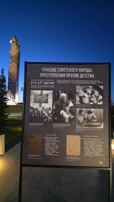 The image shows an outdoor information sign in Zaitsevo (Verevskoe S/P), Russia. The sign details the "Genocide of the Soviet People: Crimes Against Childhood" and cites that 216,431 children were killed by the Nazis in the USSR. It includes black-and-white photographs and historical documents, with text in Russian. In the background, a statue and illuminated structures are partially visible against a dusk or night sky.