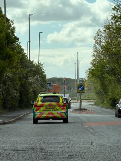 The image depicts an ambulance, registration number KX22 JWO, stopped at a traffic light in South Tyneside, United Kingdom. The traffic light is displaying a red signal. A blue and yellow road sign indicating a sharp turn is also visible. The surrounding environment includes trees, street lamps, and a road with other vehicles, suggesting a roadside location. The sky is cloudy.