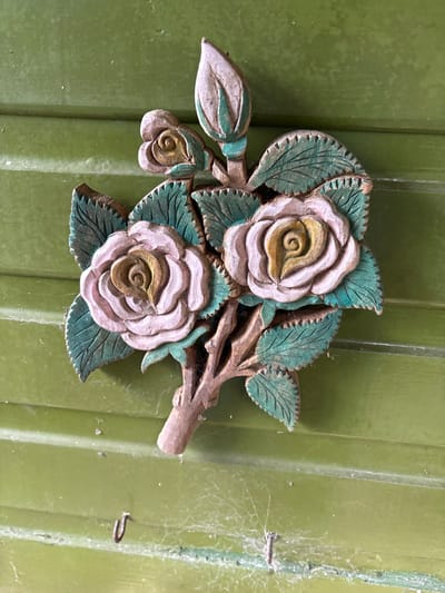 A carved wooden floral decoration, depicting a rose branch with three open roses and one rosebud, is affixed to a vertically paneled, muted green wooden surface. The roses feature pale pink-lilac outer petals with dull yellow-ochre centers. The leaves are painted a dark teal-green with visible veining. The stem is brown, textured to resemble wood. The green wooden background exhibits distinct horizontal panel lines. Cobwebs are present across the lower section of the green surface, obscuring two small, dark, U-shaped hooks located beneath the carving. The scene is situated in Stiperstones, United Kingdom.