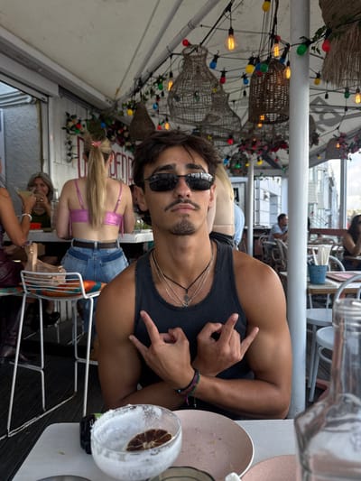 The scene depicts an open-air dining establishment located in Bondi Beach, Australia. A male subject, appearing to be in his 20s, with dark hair, a mustache, and a small goatee, is centered in the foreground, looking directly at the camera. He wears dark Ray-Ban sunglasses, a dark grey ribbed tank top, three layered necklaces (two silver chains, one black cord with a pendant), and a multi-colored string bracelet on his right wrist. Both his hands are raised, performing a "devil horns" gesture. On the white table in front of him, a glass with a white, frothy residue and a dried citrus slice is present alongside a light pink plate. A clear glass bottle is partially visible on the far right.

In the mid-ground, a female with blonde hair in a high ponytail, wearing a shiny pink crop top and blue jeans, is seated with her back to the camera on a white stool with a multi-colored cushion. To her left, a female with light brown hair holds a drink and smiles. The ceiling overhead features a white awning adorned with numerous colorful string lights, larger Edison-bulb lights, and several large, rustic woven light fixtures. White walls with partial red lettering and floral decorations are visible in the background, along with blurred figures of other patrons and residential buildings. A white structural pillar is present on the right side of the frame.