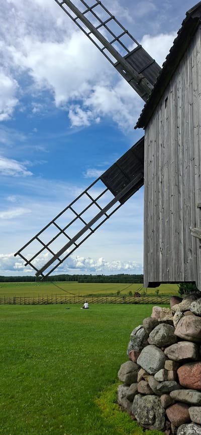 In Angla, Estonia, a weathered wooden windmill stands on a grassy field. The windmill's large, wooden blades extend diagonally across the frame against a bright blue sky dotted with white clouds. The main body of the windmill, also constructed of weathered wood planks, is visible on the right side of the image. In the distance, a fence encloses a field of green crops, with scattered hay bales visible. A single person, wearing a white shirt and dark pants, is sitting in the grass in the middle ground, facing away from the camera. In the foreground, a pile of large, rounded stones creates a natural border on the right.