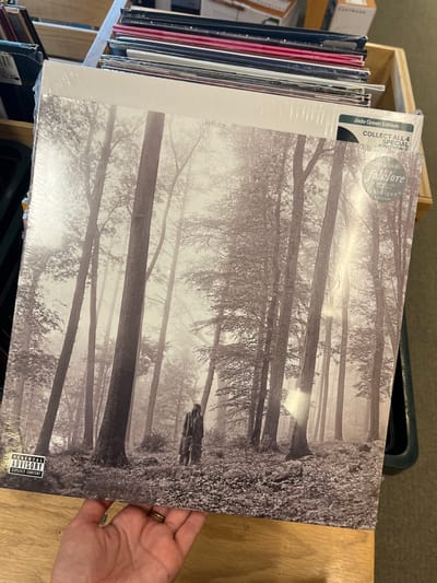 A sealed vinyl record album, "folklore" by Taylor Swift, "Jade Green Edition," is depicted held by a human hand with light skin and a ring on the fourth finger. The album cover presents a grayscale image of Taylor Swift in a long coat, standing among numerous tall, slender-trunked trees in a misty forest. A "PARENTAL ADVISORY EXPLICIT CONTENT" label is visible in the lower-left corner of the cover. Additional labels include "Taylor Swift folklore" and "cardigan" on a circular sticker, and "Jade Green Edition" with "COLLECT ALL 4 SPECIAL EDITIONS" on a rectangular sticker in the upper-right. The album is held above a light-colored wooden surface. In the background, a stack of multiple other vinyl records is arranged vertically in a display rack or bin, partially obscured. A dark green container is visible to the far left. The specific location within the city of El Paso, United States, is not discernible from the provided image.