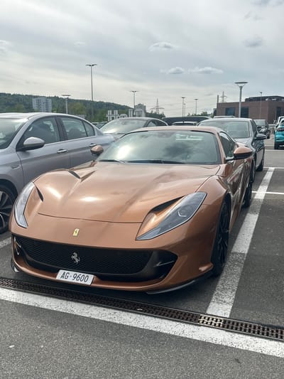 The image, taken in Wallisellen, Switzerland, shows a distinctive bronze or gold Ferrari parked in an asphalt parking lot during the daytime under a partly cloudy sky. The car is the primary subject, viewed from the front-right quarter, showcasing its sleek design, prominent front grille, and modern headlights. The Ferrari emblem is visible on the grille.
To the left of the Ferrari, other cars are parked, including a grey sedan. The parking lot features white painted lines and a metal drainage grate running alongside the Ferrari. In the background, there are several utility poles or streetlights, power lines, distant buildings, and a line of trees suggesting a wooded area or distant landscape. The sky is bright, with some cloud formations, and reflections on the Ferrari's bodywork indicate sunlight.
No people are visible in the image, and the cars appear to be stationary. The visible text on the license plate of the Ferrari is "AG-9600." The overall impression is of a luxury sports car parked in an urban or suburban setting.